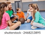 A group of diverse children sit on the floor listening to a male teacher instructions, a basketball in the center.