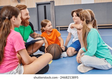 A group of diverse children sit on the floor listening to a male teacher instructions, a basketball in the center. - Powered by Shutterstock