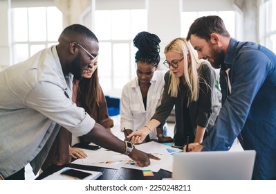 Group of diverse colleagues in casual clothes gathering around table with laptop smartphone and discussing details of project while showing with fingers at documents content and working in office - Shutterstock ID 2250248211