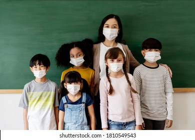 Group Of Diverse Elementary Students In Classroom Alongside With Young Adult Asian Teacher. Primary School Multi-racial Pupils Standing In A Row Wearing A Face Mask. School During Covid19 Concept