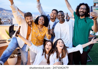 Group of diverse excited friends celebrating with beers at barbecue party in attic. Portrait of young people posing raising arms on terrace in evening in open air. Cheerful colleagues enjoying time. - Powered by Shutterstock