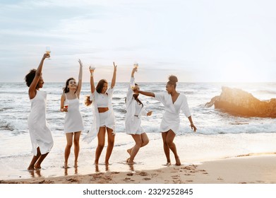 Group of diverse female friends drinking champagne while dancing and having hen party on the beach, copy space. Women celebrating bachelorette party during summer sunset - Powered by Shutterstock