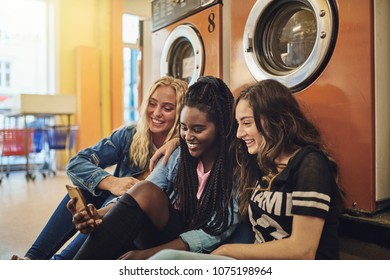 Group Of Diverse Female Friends Laughing Together While Sitting On The Floor Of A Laundromat Using A Cellphone