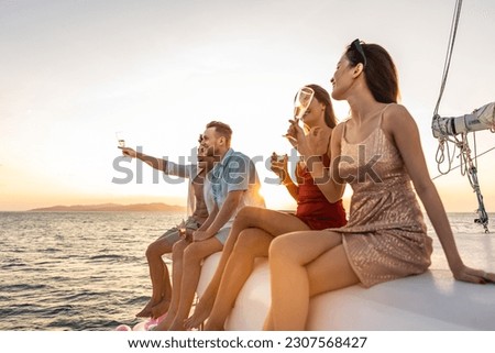 Group of diverse friend sit on deck of yacht while yachting together. Attractive young men and women hanging out, celebrating holiday vacation trip while catamaran boat sailing during summer sunset.