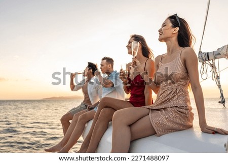 Group of diverse friends drink champagne while having a party in yacht. Attractive young men and women hanging out, celebrating holiday vacation trip while catamaran boat sailing during summer sunset.