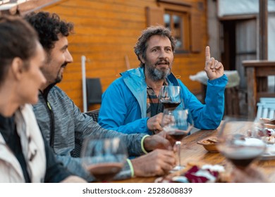 Group of diverse friends engaged in animated conversation during outdoor wine tasting. Casual atmosphere with rustic wooden table, red wine glasses, and lively discussion - Powered by Shutterstock