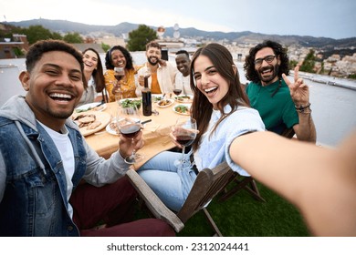 Group of diverse friends having fun at rooftop party. Beautiful woman taking selfie at barbecue dinner time. Smiling people eating and drinking on outdoor terrace. Positive friendship relations. - Powered by Shutterstock