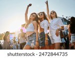 Group of diverse girls having fun at coastal music festival. Women dance, smile in sunlight by sea. Friends enjoy live band, sunset party vibes on sandy beach. Youth culture, summer event gathering.