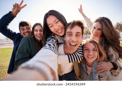 Group of diverse happy young friend taking selfie together looking smiling at camera. Millennial people sharing cheerful moments posing piggyback with peace gestures raised. Friendship and community - Powered by Shutterstock