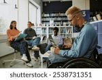 Group of diverse individuals engaging in study session in modern library workspace. Man in wheelchair focusing on work with laptop, while others discussing and taking notes