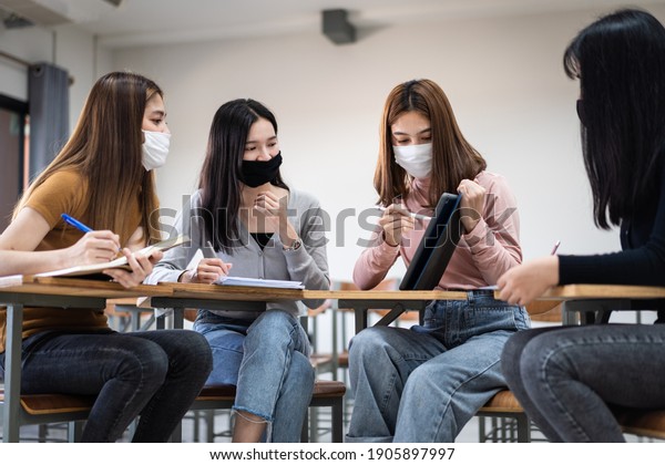 Group of diverse international students\
wearing protective  masks and talking, discussing project, sitting\
at desk in the classroom at the\
university