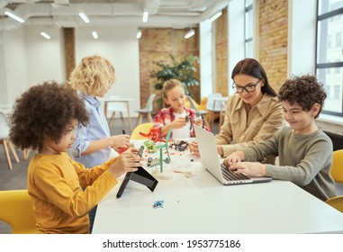 Group of diverse kids working together with young female teacher, using digital devices and playing with mechanical toys while sitting at the table during STEM class