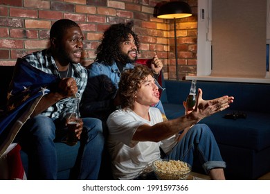 Group Of Diverse Men Sitting On Sofa Watching Sport Championship Together Emotionally Reacting On Game Process, Gesturing, Drinking Beer, At Home