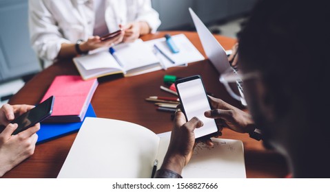 Group of diverse modern employees browsing mobile phones and laptop sitting at table loaded with notepad and stationery in modern office
