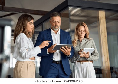 Group of diverse partners mature Latin business man and European business women discussing project on tablet in office. Team of colleagues professionals business people working together at workspace. - Powered by Shutterstock