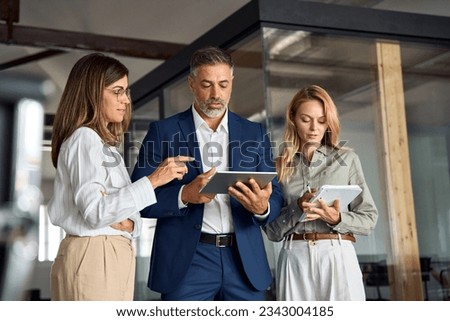 Group of diverse partners mature Latin business man and European business women discussing project on tablet in office. Team of colleagues professionals business people working together at workspace.
