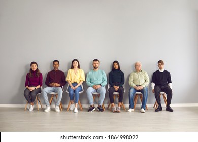 Group Of Diverse People In Casual Clothes Waiting In Line To See Doctor. Male And Female Candidates Of Different Ages And Ethnicities Sitting On Chairs In Row In Office Corridor Before Job Interview