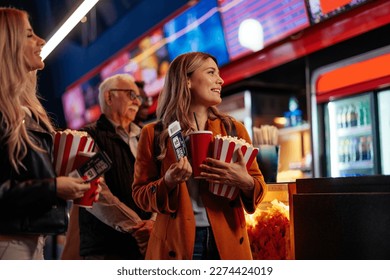 A group of diverse people are queueing up in a movie theater to watch a movie. They are holding their tickets, popcorn and drinks in their hands.