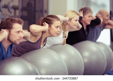 Group of diverse people in a receding line doing pilates in a gym balancing over the gym balls with their hands laocked behind their necks toning their muscles