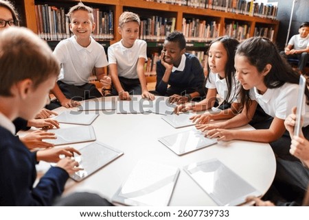 Group of diverse students in a library using tablets. Kids in school uniforms engaged in learning. Education and technology in a classroom setting. Diverse elementary school kids in library.