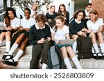 Group of diverse students in school uniforms sitting on steps. Students using laptops and phones. Boys and girls in uniforms. School uniforms and technology. Diverse teenage students at school.