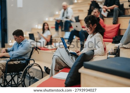 A group of diverse students study and take notes in a tiered lecture space. Several learners use laptops and notebooks while one student in a wheelchair participates