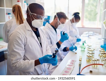 Group of diverse technicians wearing face masks and coats working together with samples at a table in a lab - Shutterstock ID 1932560591
