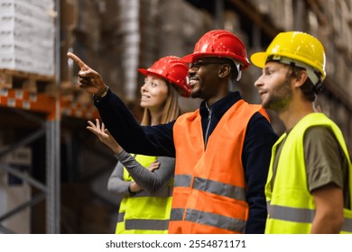 A group of diverse warehouse workers wearing safety gear, smiling and discussing logistics. The team is focused and engaged, showcasing teamwork and collaboration in an industrial setting. - Powered by Shutterstock