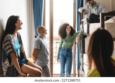 A group of diverse young adults relax in a hostel dorm room, laughing and chatting together. One individual has a shaved head, piercings, and androgynous appearance - diversity and people lifestyle
