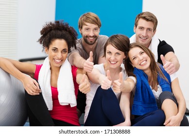 Group of diverse young athletes at the gym sitting on stairs giving a thumbs up of approval as they smile happily at the camera - Powered by Shutterstock