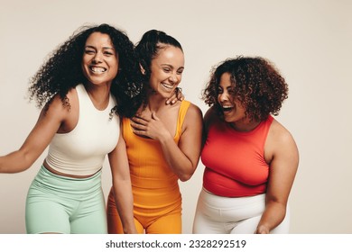 Group of diverse young female athletes are smiling and laughing as they stand together together in the studio dressed in sportswear, celebrating their healthy lifestyle and friendship.