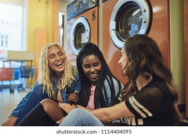 Group Of Diverse Young Female Friends Sitting On A Laundromat Floor Talking And Laughing Together While Doing Laundry