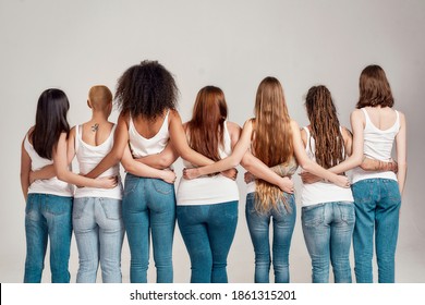 Group of diverse young women wearing white shirt and denim jeans putting arms around each other while posing together isolated over grey background. Diversity, body positivity, friendship. Back view