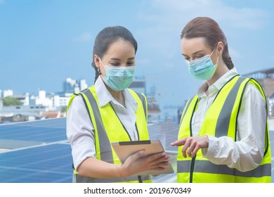 Group Of Diversity Factory Worker Women With Face Mask Meeting To Inspect At Rooftop Of Solar Cell Panel In Background. Asian, And White Caucasian People Operate On Site Renewable Energy Construction.