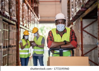 Group Of Diversity Workers Wearing Protective Face Mask Working In Factory Warehouse. Black Man Pushing Metal Lathe Cart With Box Parcel During Covid 19 Pandemic Crisis. Logistic Industry Concept.