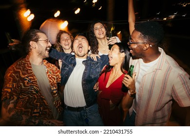 A group of drunk multiracial friends is having fun at the night open-air party. They are singing, drinking beer, and having a good time together.
