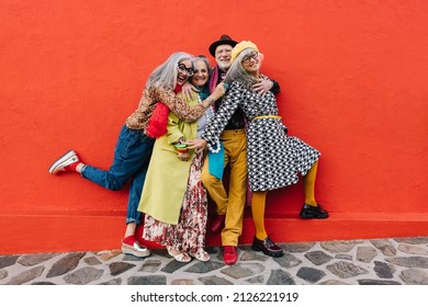 Group Of Eccentric Senior Citizens Laughing And Having Fun While Standing Together Against A Red Background. Four Cheerful Elderly Friends Celebrating Their Retirement In Colourful Clothing.