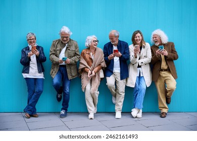 Group of elderly Caucasian laughing together using cell phone standing leaning on blue wall. Happy old friends with gray hair gathered having fun looking mobile outdoor. Mature people enjoying devices - Powered by Shutterstock