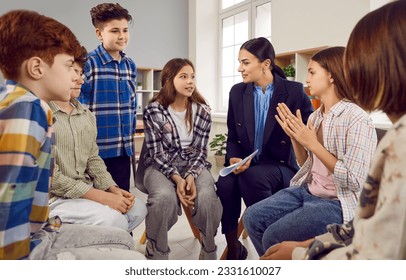 Group of elementary school children sitting in circle during class. Boys and girls students in casual wear sitting on chairs and talking to friendly female teacher in classroom - Powered by Shutterstock