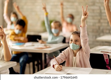 Group Of Elementary Students With Face Masks                       Raising Hands To Answer The Teacher's Question In The Classroom. Focus Is On A Girl. 