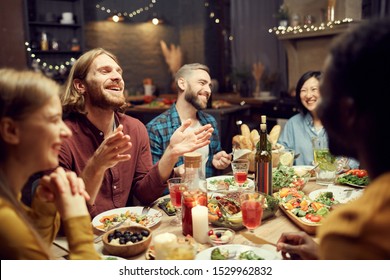 Group Of Emotional Young People Enjoying Dinner Party With Friends And Smiling Happily Sitting At Table In Dimly Lit Room, Copy Space