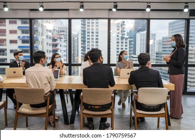 Group of employees sitting at a meeting, wearing a mask, having a leader standing at the head of the table in the meeting, introducing new normal work.