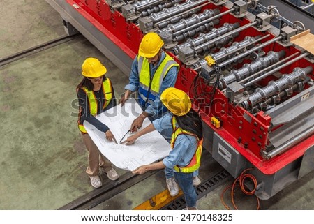 A group of engineers from different races and ethnicities working closely together using a blueprint plan to design or control a manufacturing system next to metal sheet maker machine.
