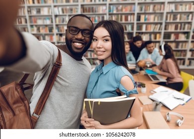 Group Of Ethnic Multicultural Students Sitting At Table In Library. Black Guy And Asian Girl Taking Selfie On Phone.