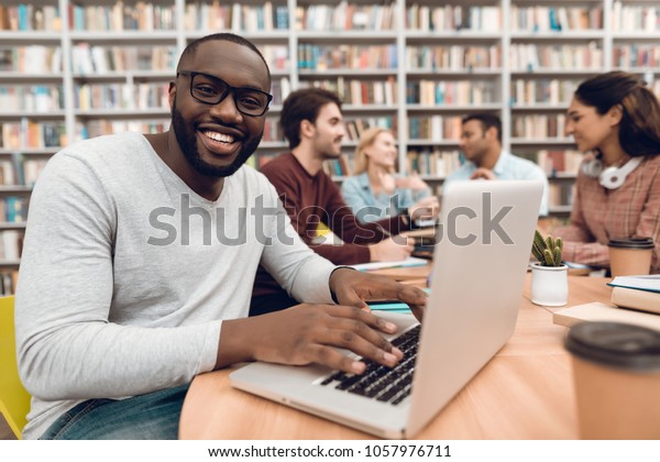 Group of ethnic multicultural students\
sitting at table in library. Black guy on\
laptop.