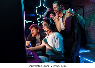 Group of excited friends celebrating victory of young asian girl in online video game while standing next to her in modern cybersport club