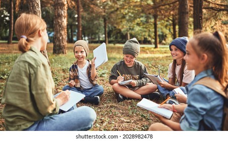 Group of excited school children boys and girls with young female teacher sitting on ground in forest during ecology lesson outdoors, writing down in notebooks their discoveries of exploring nature - Powered by Shutterstock