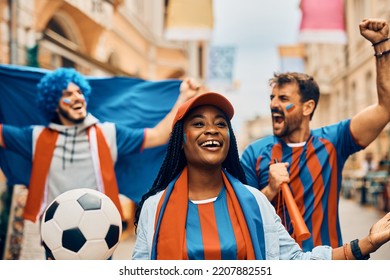 Group Of Excited Soccer Fans Cheering While Walking On The Street. Focus Is On African American Woman. 