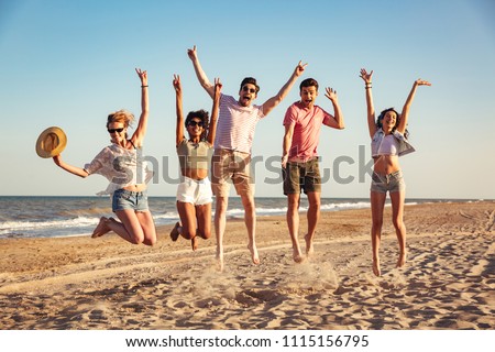 Group of excited young friends dressed in summer clothes holding hands while jumping together at he beach