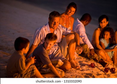 Group Of Family And Friends Sitting In The Glow Of A Campfire On A Sandy Beach At Night.